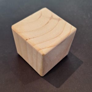 May include: A light-coloured wooden cube with rounded edges. The wood grain is visible on the top surface. The cube is sitting on a dark surface, casting a shadow.