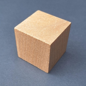 May include: A light brown wooden cube, displaying the natural grain of the wood. The cube sits on a dark grey surface, emphasizing its geometric shape and texture. The precisely cut edges of the cube highlight its simple, yet elegant design.