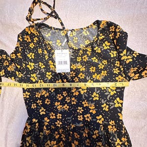 May include: Black dress with yellow floral pattern. The dress has long sleeves and a tie at the neckline. A measuring tape shows the dress is approximately 18 inches long. The tag says "Free People".