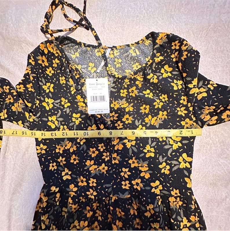 May include: Black dress with yellow floral pattern. The dress has long sleeves and a tie at the neckline. A measuring tape shows the dress is approximately 18 inches long. The tag says "Free People".