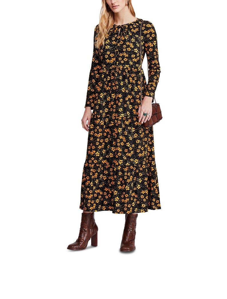 May include: A long-sleeved black dress with a yellow floral pattern. The dress has a tiered skirt and a tie detail at the neckline. The model is wearing brown ankle boots.