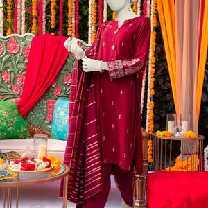 Puede incluir: Un salwar kameez de color marrón con bordados dorados y un dupatta a juego. El atuendo lo lleva un maniquí y se exhibe sobre un fondo de decoraciones florales rojas y naranjas.