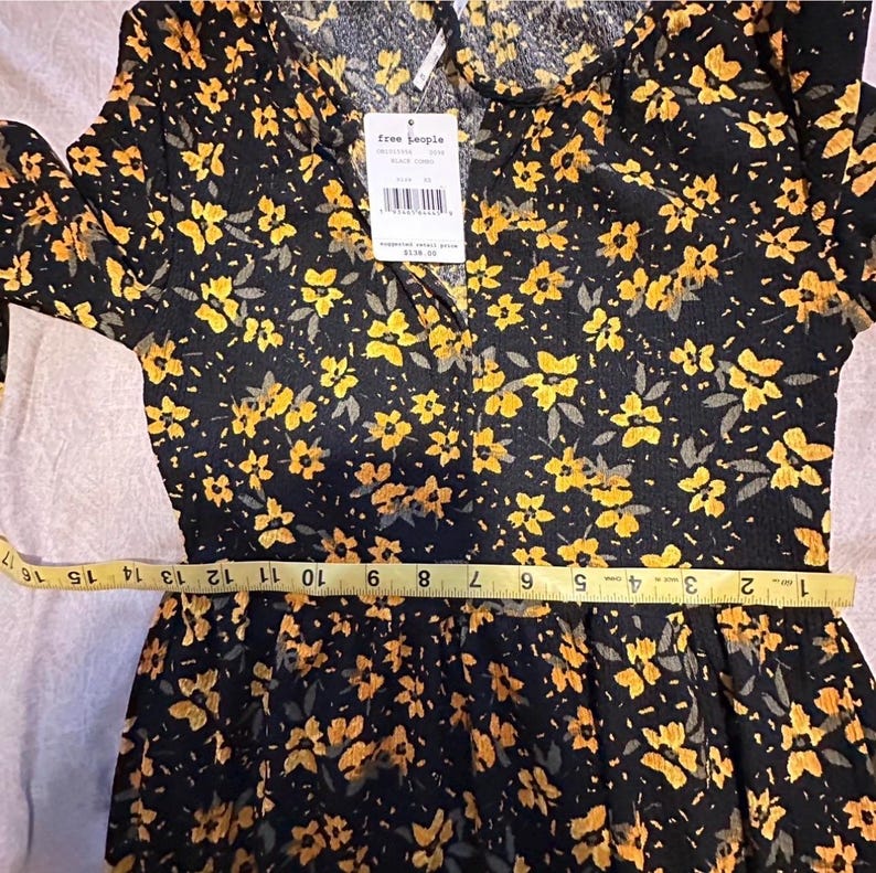 May include: Black dress with a yellow floral pattern. The dress has long sleeves and a collared neckline. A measuring tape is placed across the waist. The tag reads "free people".