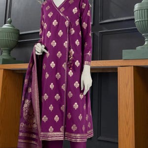 May include: A purple and gold patterned dress with a long-sleeved top and matching pants. The dress has a V-neckline and a gold tassel detail. The pants have a wide leg and a gold trim. The dress is paired with a purple and gold patterned dupatta.
