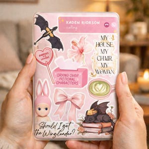 May include: A pink phone case decorated with stickers, including a dragon, a bow, and the text "Should I get the Winglender?" and "Crying over fictional characters." The case is held in two hands.