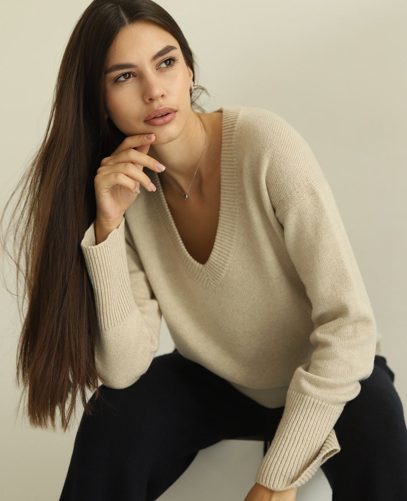 Puede incluir: Un su&eacute;ter beige de manga larga con un cuello en V profundo y pu&ntilde;os anchos acanalados. La prenda est&aacute; confeccionada en un material de punto suave y se combina con pantalones azul marino oscuro, creando un estilo neutro para el armario.