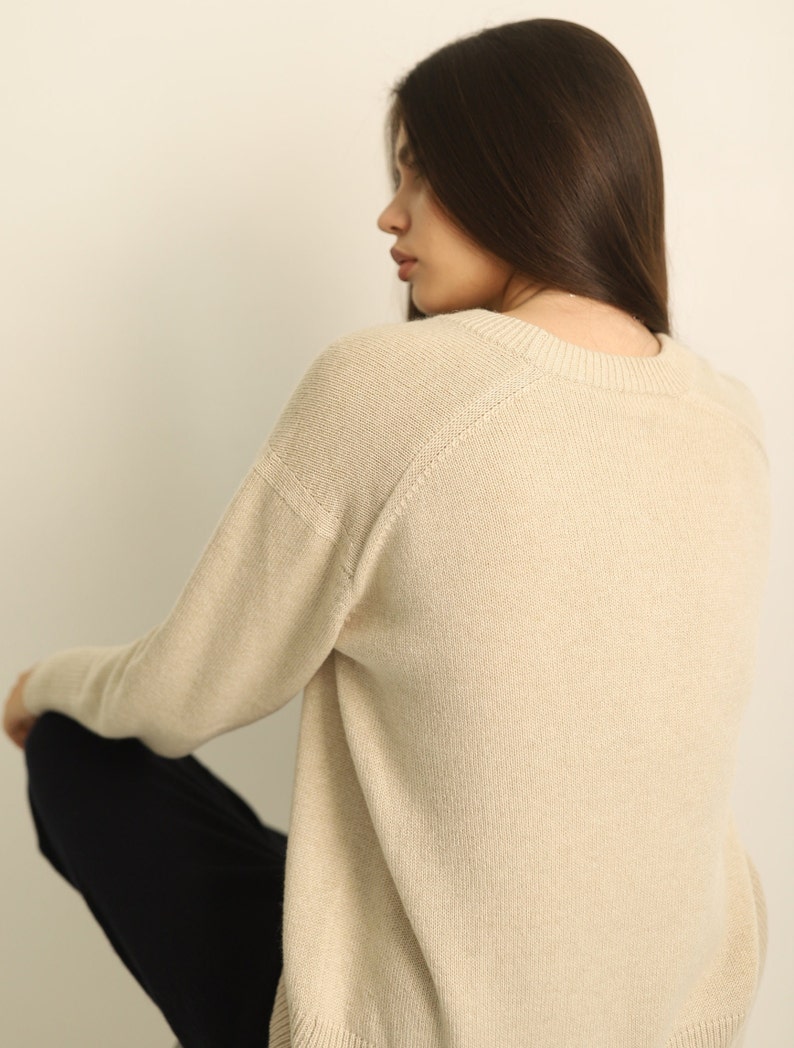 Puede incluir: Un su&eacute;ter beige de manga larga con cuello redondo y mangas ragl&aacute;n. La prenda de punto presenta un cuello, pu&ntilde;os y dobladillo acanalados, mostrando una textura lisa y de color s&oacute;lido en toda la parte posterior y el perfil lateral.