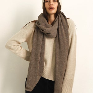 Puede incluir: Un gorro de punto color crema y una bufanda larga de color marr&oacute;n s&oacute;lido sobre un su&eacute;ter de cuello en V color crema con pu&ntilde;os y dobladillo acanalados, combinado con pantalones negros. El conjunto presenta un look en capas de tonos neutros.