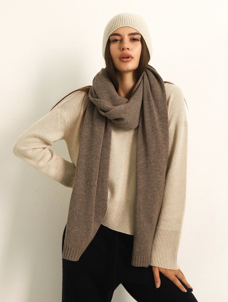 Puede incluir: Un gorro de punto color crema y una bufanda larga de color marr&oacute;n s&oacute;lido sobre un su&eacute;ter de cuello en V color crema con pu&ntilde;os y dobladillo acanalados, combinado con pantalones negros. El conjunto presenta un look en capas de tonos neutros.