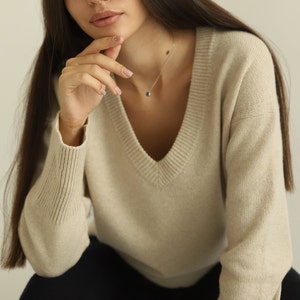 Puede incluir: Un su&eacute;ter beige con cuello en V, mangas largas con pu&ntilde;os acanalados y cuello acanalado. El tejido de punto tiene una textura jaspeada sutil. Un delicado collar de cadena plateada con un peque&ntilde;o colgante redondo oscuro descansa en el cuello.