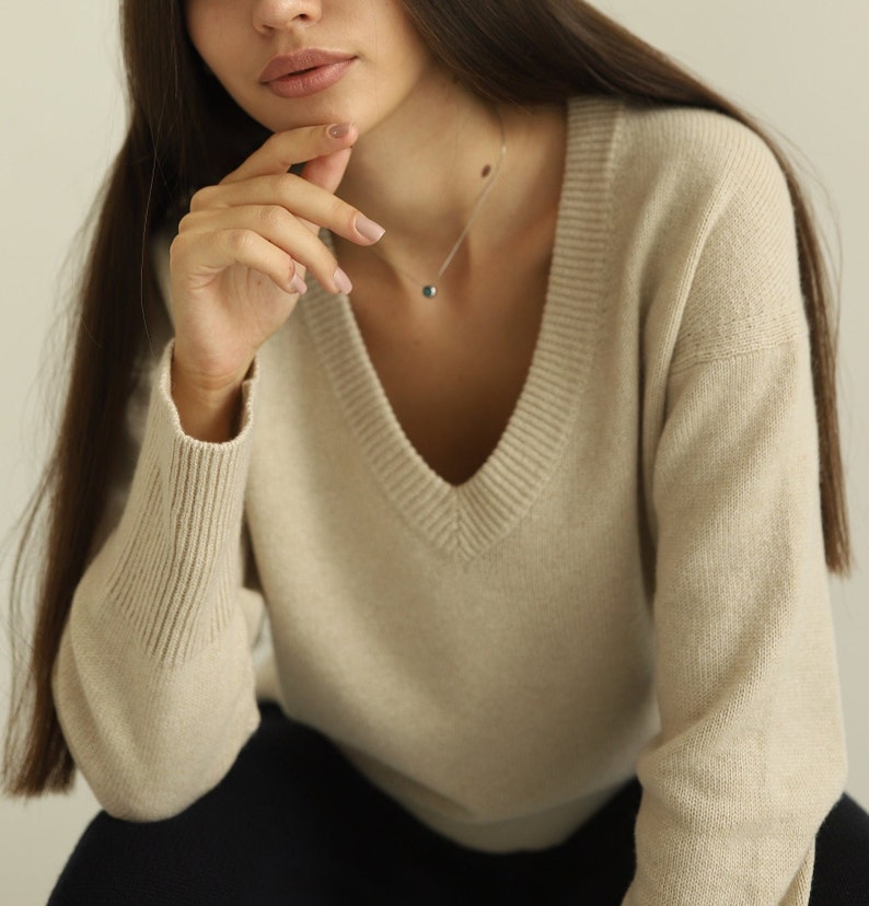 Puede incluir: Un su&eacute;ter beige con cuello en V, mangas largas con pu&ntilde;os acanalados y cuello acanalado. El tejido de punto tiene una textura jaspeada sutil. Un delicado collar de cadena plateada con un peque&ntilde;o colgante redondo oscuro descansa en el cuello.