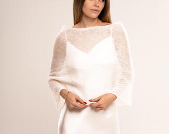May include: White knitted shrug with a loose, open weave. The shrug has a boat neck and long sleeves. It is shown draped over a white slip dress.