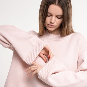 Puede incluir: Un suéter suave de color rosa pálido con mangas largas y un ajuste holgado. El suéter tiene un cuello redondo y una longitud ligeramente recortada.