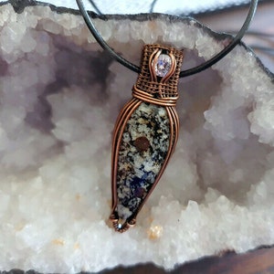 May include: A copper wire wrapped pendant featuring a speckled grey and black stone with a small purple gemstone at the top.