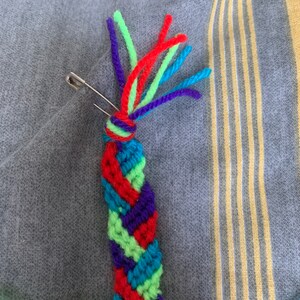 May include: A colourful friendship bracelet being made with green, red, blue and purple yarn. A safety pin is holding the yarn in place.