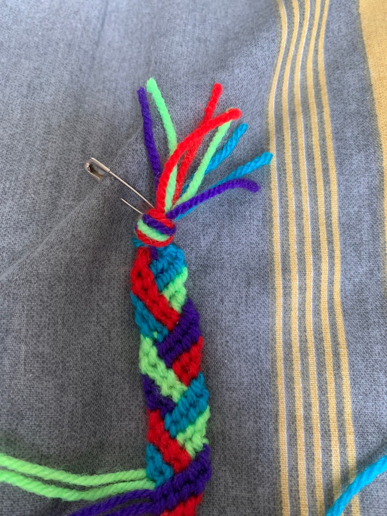 May include: A colourful friendship bracelet being made with green, red, blue and purple yarn. A safety pin is holding the yarn in place.