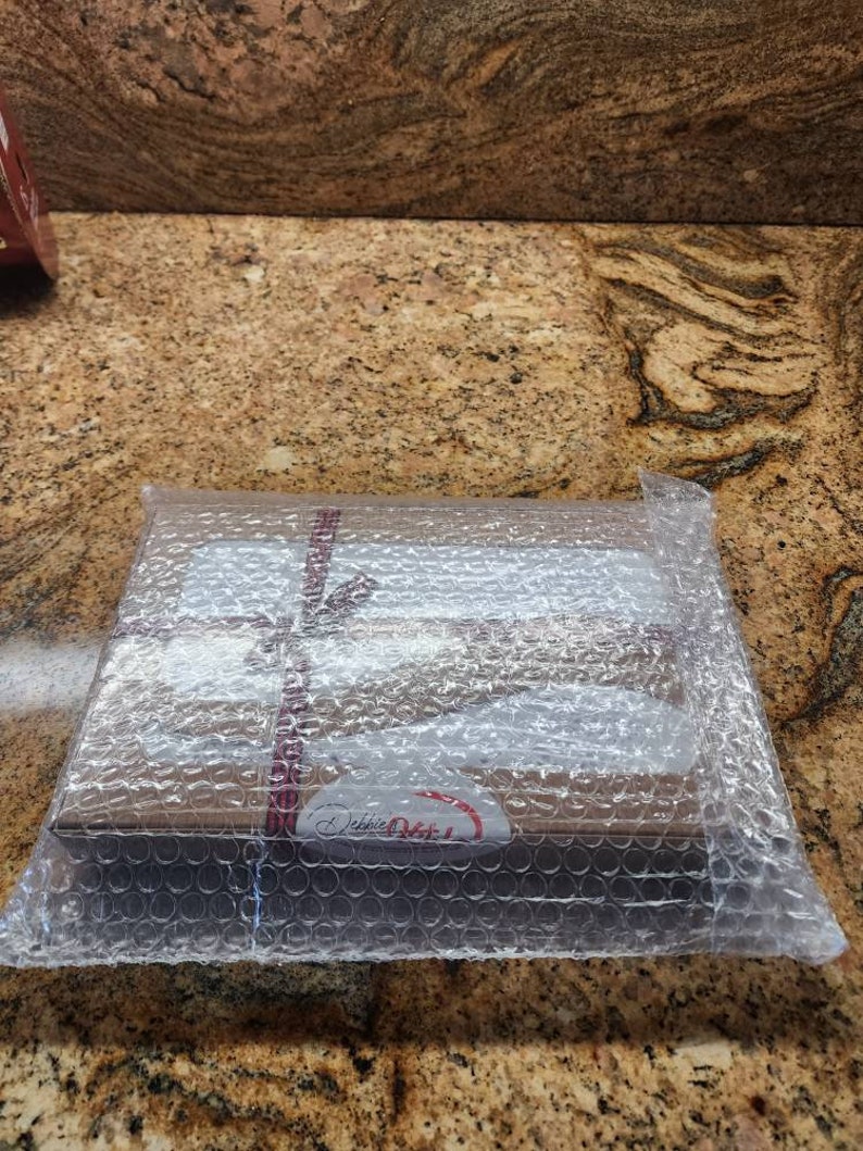 Peut inclure: Une bo&icirc;te rectangulaire en carton brun envelopp&eacute;e dans du papier bulle transparent. La bo&icirc;te est fix&eacute;e par un fin ruban &agrave; motifs rouges et blancs nou&eacute; en boucle. Une &eacute;tiquette ovale blanche avec le texte Debbie Meyer est visible sur le devant.