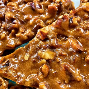 Peut inclure: Une vue en gros plan de morceaux de croquant aux noix de p&eacute;can, pr&eacute;sentant une base de sucre caram&eacute;lis&eacute; ambr&eacute; et brillant, incrust&eacute;e de nombreux morceaux de noix de p&eacute;can bruns hach&eacute;s, cr&eacute;ant une surface textur&eacute;e et in&eacute;gale &agrave; l'aspect collant.