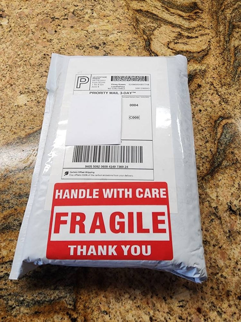 Peut inclure: Une enveloppe d'exp&eacute;dition rectangulaire blanche avec une &eacute;tiquette d'exp&eacute;dition Priority Mail 3-Day comprenant un code-barres et un num&eacute;ro de suivi. Un grand autocollant rectangulaire rouge porte le texte blanc HANDLE WITH CARE FRAGILE THANK YOU.