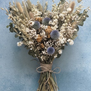 Puede incluir: Un ramo de flores secas con una est&eacute;tica r&uacute;stica y natural. Presenta una mezcla de hierbas secas, flores blancas y cardos azules, atados con cuerda.