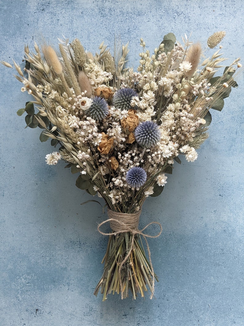 Puede incluir: Un ramo de flores secas con una est&eacute;tica r&uacute;stica y natural. Presenta una mezcla de hierbas secas, flores blancas y cardos azules, atados con cuerda.