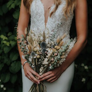 Puede incluir: Una novia sostiene un ramo de flores secas en tonos beige, verde y azul. El ramo incluye hierbas secas, eucalipto y peque&ntilde;as flores de cardo azul.