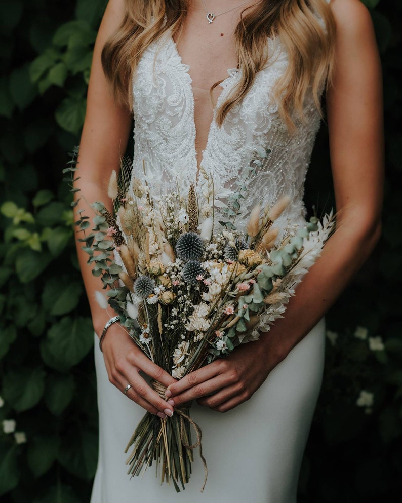 Puede incluir: Una novia sostiene un ramo de flores secas en tonos beige, verde y azul. El ramo incluye hierbas secas, eucalipto y peque&ntilde;as flores de cardo azul.