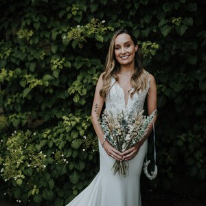 Puede incluir: Una novia con un vestido de novia blanco con una larga cola, sosteniendo un ramo de flores secas. Est&aacute; de pie frente a una pared de hiedra verde.