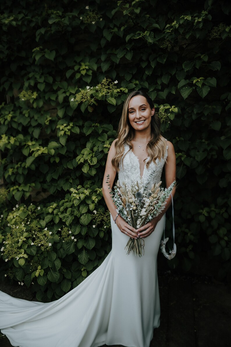 Puede incluir: Una novia con un vestido de novia blanco con una larga cola, sosteniendo un ramo de flores secas. Est&aacute; de pie frente a una pared de hiedra verde.