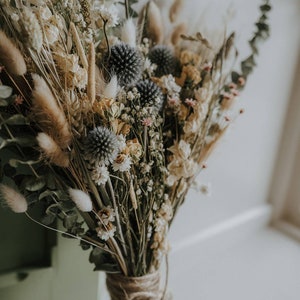 Puede incluir: Un ramo de flores secas con una variedad de flores beige, blancas y grises, incluyendo hierbas secas y hojas de eucalipto. El ramo est&aacute; atado con cuerda.