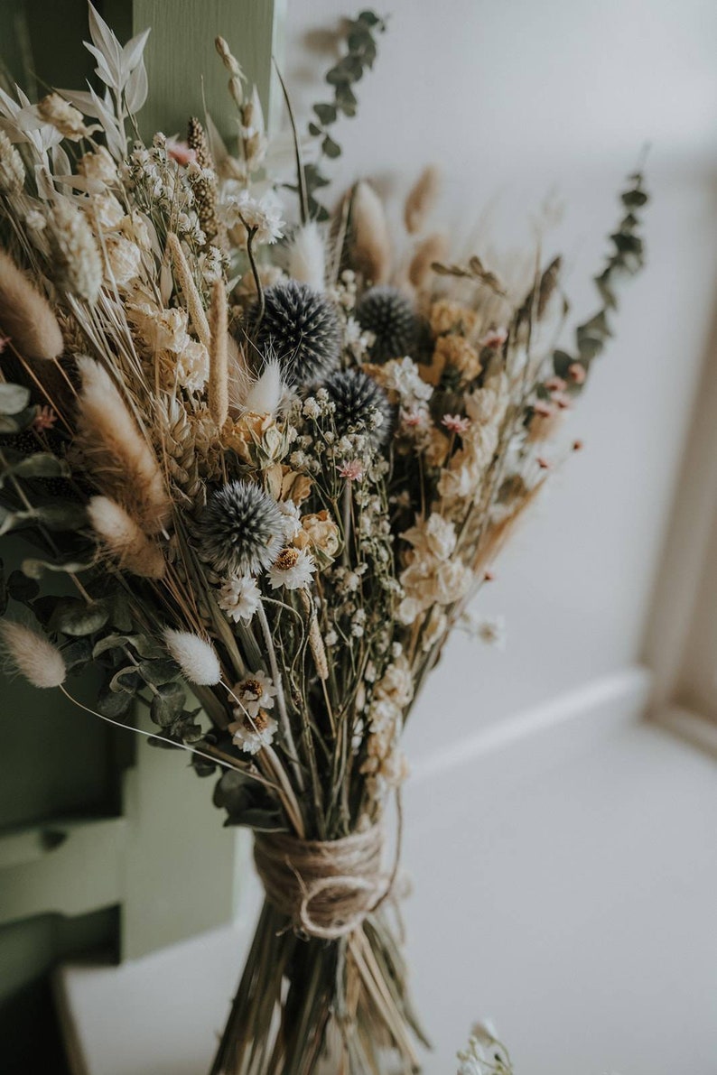 Puede incluir: Un ramo de flores secas con una variedad de flores beige, blancas y grises, incluyendo hierbas secas y hojas de eucalipto. El ramo est&aacute; atado con cuerda.