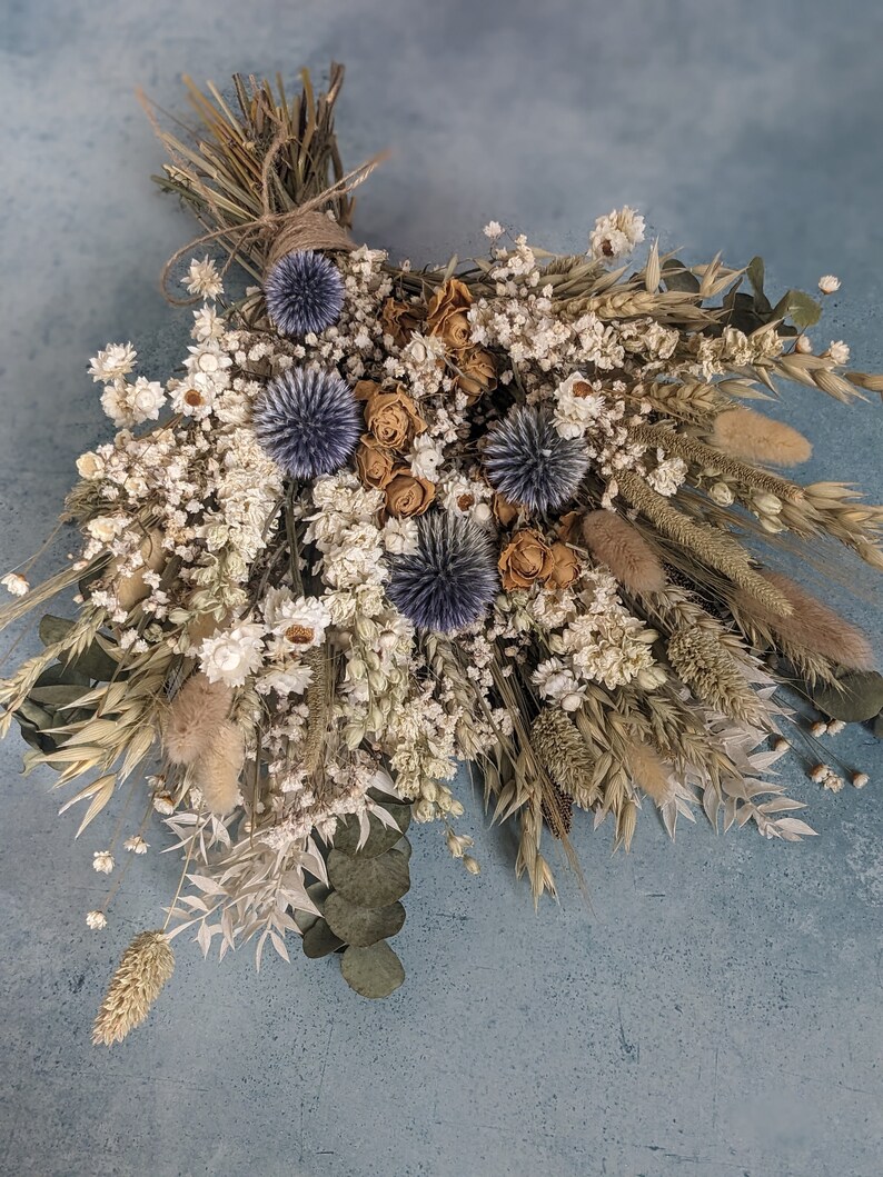 Puede incluir: Un ramo de flores secas con flores blancas, beige y azules. El ramo est&aacute; atado con cuerda e incluye hierbas secas y eucalipto.