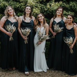 Puede incluir: Cinco mujeres en fila, vestidas con vestidos de sat&eacute;n negro y sosteniendo ramos de flores secas. La mujer del centro lleva un vestido de novia blanco.