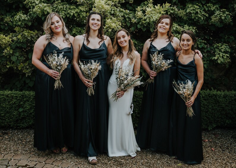 Puede incluir: Cinco mujeres en fila, vestidas con vestidos de sat&eacute;n negro y sosteniendo ramos de flores secas. La mujer del centro lleva un vestido de novia blanco.