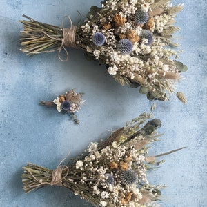 Puede incluir: Dos ramos de flores secas con una est&eacute;tica r&uacute;stica y natural. El ramo m&aacute;s grande presenta una variedad de hierbas secas, flores blancas y cardos azules. El ramo m&aacute;s peque&ntilde;o es una composici&oacute;n similar pero de menor tama&ntilde;o.
