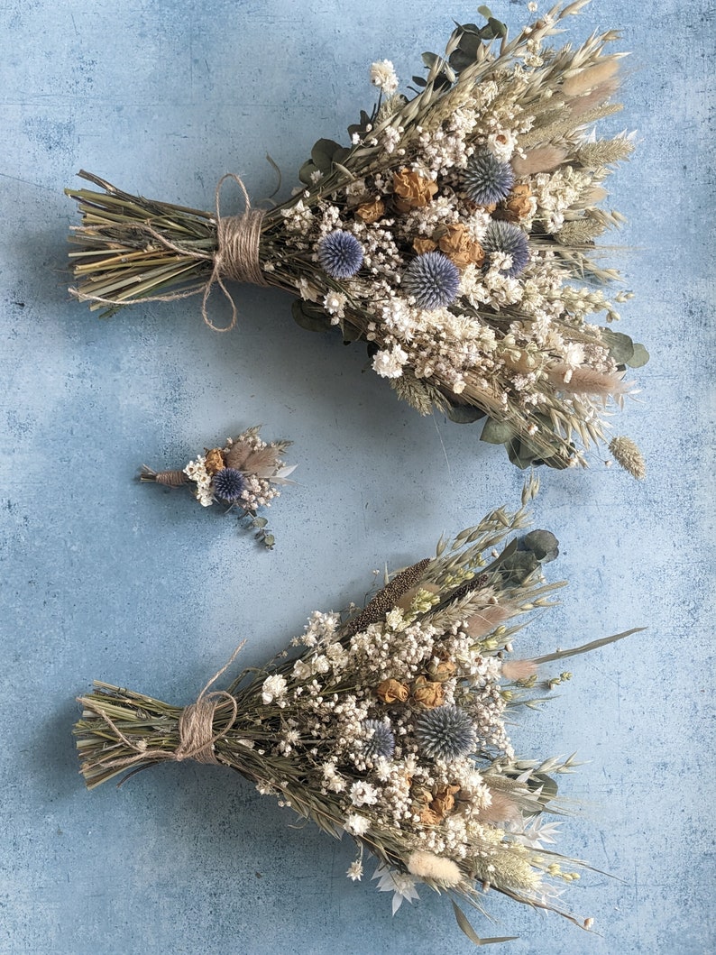 Puede incluir: Dos ramos de flores secas con una est&eacute;tica r&uacute;stica y natural. El ramo m&aacute;s grande presenta una variedad de hierbas secas, flores blancas y cardos azules. El ramo m&aacute;s peque&ntilde;o es una composici&oacute;n similar pero de menor tama&ntilde;o.