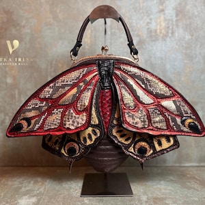 Può includere: Una borsa a forma di falena con manico marrone scuro e chiusura in metallo. Le ali presentano motivi strutturati nei toni del rosso, oro, nero e grigio. Il testo VITKA IRINA DESIGNER BAGS è visibile nell'angolo in alto a sinistra dell'immagine.