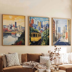 Op de afbeelding: Drie ingelijste kunstprints met stadsgezichten hangen boven een bruine bank. De kunst toont schilderijen van stadsgezichten in aquarel, waaronder een brug, een gele tram en een straatbeeld. De kamer is ingericht met kussens, een ronde houten salontafel en een geweven vloerkleed.