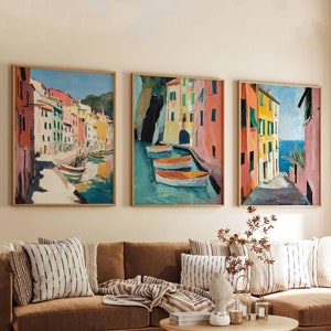 May include: Three framed paintings of colorful buildings and boats in a harbor. The paintings are hung on a wall above a brown sectional sofa with throw pillows and a blanket.