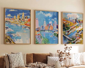Pode incluir: Três pinturas emolduradas de paisagens urbanas em tons de azul e amarelo adornam uma sala de estar. As obras de arte retratam edifícios e pontes. Um sofá castanho, uma mesa de centro redonda de madeira e um tapete tecido completam a cena.