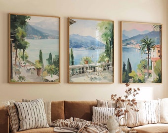 Puede incluir: Tres acuarelas enmarcadas que representan paisajes pintorescos con agua, árboles y edificios. Las pinturas están colgadas en una pared sobre un sofá seccional marrón con almohadas y una manta.