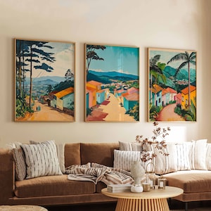 May include: Three framed landscape paintings of colourful houses and palm trees hang on a wall above a brown sectional sofa with throw pillows and a blanket.