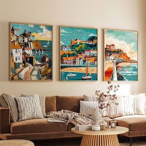 May include: Three framed paintings depicting seaside towns with colourful buildings, blue water, and boats. The paintings are hung on a wall above a brown sectional sofa with cushions and a throw blanket.