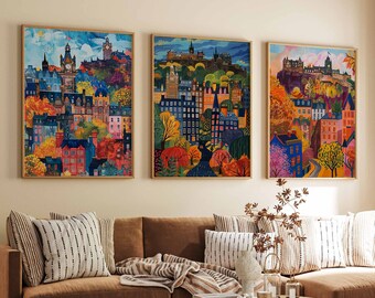 May include: Three colorful paintings of a city skyline with buildings and trees. The paintings are framed in gold and hung on a beige wall above a brown couch with pillows.