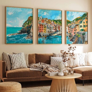 May include: Three framed paintings depicting colourful buildings on a cliff overlooking a blue ocean. The paintings are hung on a beige wall above a brown sectional sofa with cushions and a coffee table.