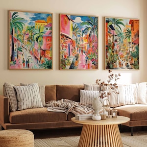 Può includere: Tre dipinti colorati che rappresentano una scena di strada cittadina con palme e edifici. I dipinti sono incorniciati in legno e appesi a una parete beige sopra un divano angolare marrone con cuscini e un tavolino da caffè.