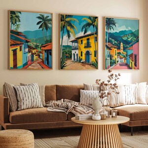 May include: Three framed colorful paintings of a tropical city with palm trees and colorful buildings. The paintings are hanging on a beige wall above a brown sectional sofa with pillows and a coffee table.