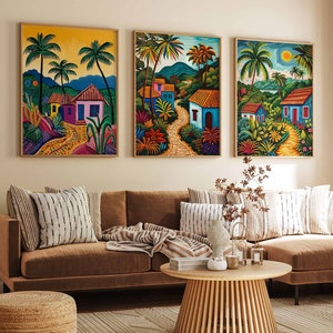 May include: Three colourful framed paintings depicting tropical scenes with houses, palm trees, and winding paths. The paintings are hung on a beige wall above a brown sectional sofa with cushions and a throw blanket.