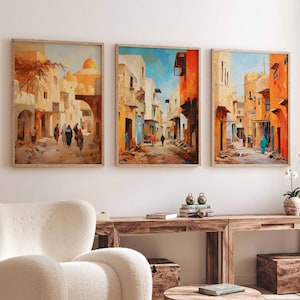 Könnte beinhalten: Drei gerahmte Bilder, die farbenfrohe Straßenszenen in einer Stadt im Nahen Osten zeigen. Die Bilder hängen an einer weißen Wand über einem Holztisch und einem weißen Sessel.