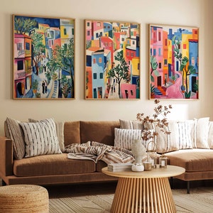 May include: Three colourful paintings depicting a city street scene with buildings, trees, and people. The paintings are framed and hung on a beige wall above a brown sectional sofa with cushions and a coffee table.